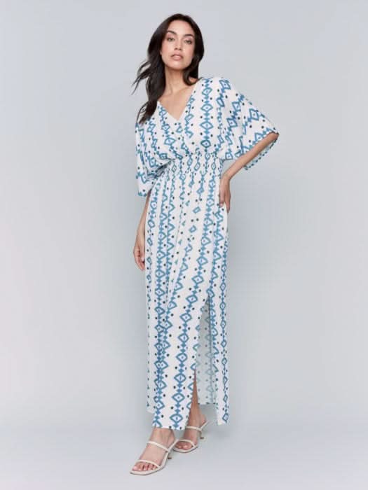 A fashionista in a Charlie B Printed Dolman Sleeve Maxi Dress with Side Slits stands sideways, eyes on the camera, hand on hip, paired with chic white heeled sandals.