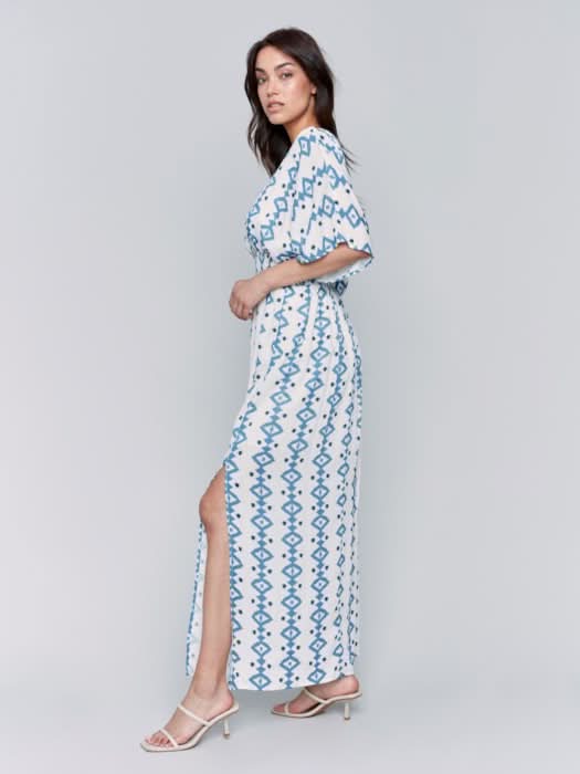A fashionista in a Charlie B Printed Dolman Sleeve Maxi Dress with Side Slits stands sideways, eyes on the camera, hand on hip, paired with chic white heeled sandals.