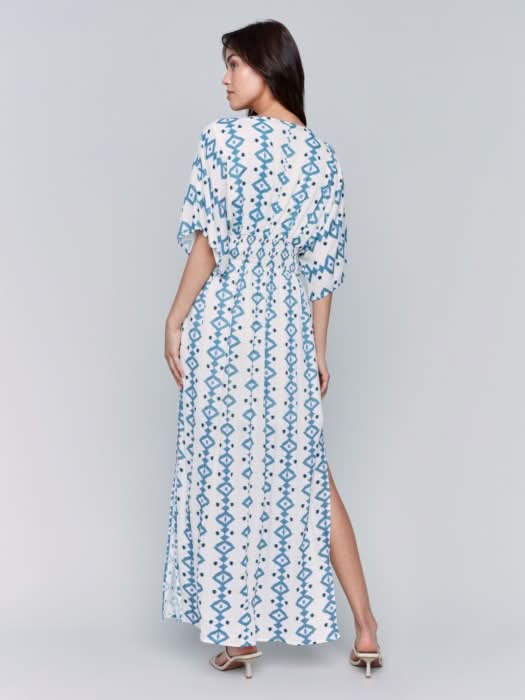 A fashionista in a Charlie B Printed Dolman Sleeve Maxi Dress with Side Slits stands sideways, eyes on the camera, hand on hip, paired with chic white heeled sandals.