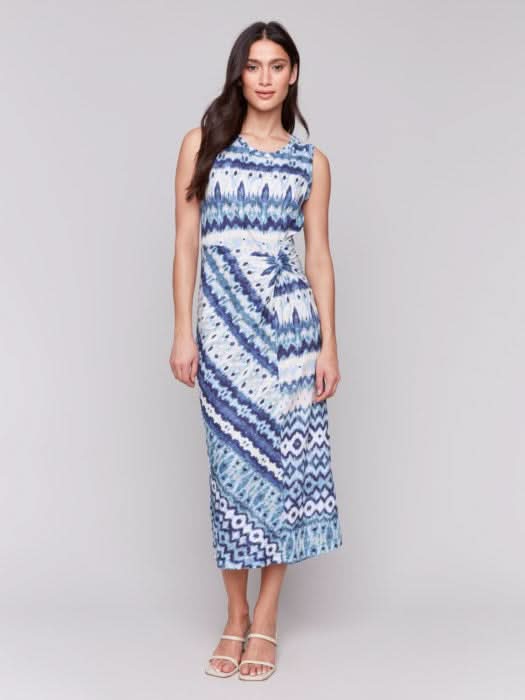 A woman stands facing away, wearing the Charlie B Sleeveless Round Neck Dress with Faux Knot Detail in a blue and white geometric pattern and heeled sandals.
