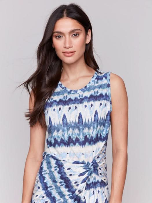 A woman stands facing away, wearing the Charlie B Sleeveless Round Neck Dress with Faux Knot Detail in a blue and white geometric pattern and heeled sandals.