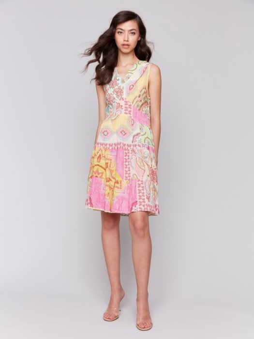 A woman models the Charlie B Sleeveless V-Neck Tiered Dress with a colorful patchwork pattern and neutral heels, embodying chic summer style against a plain background.