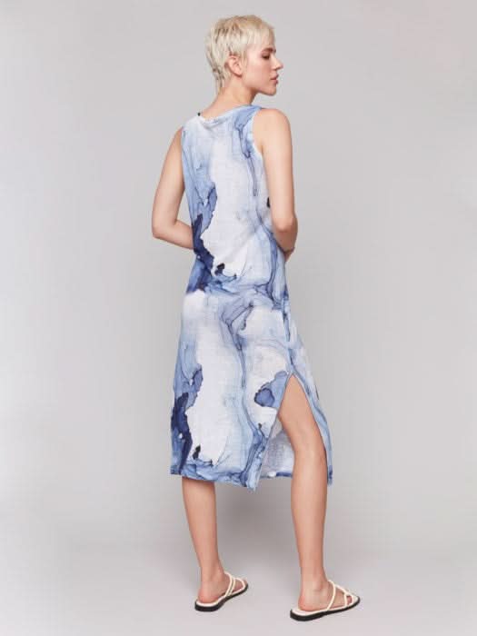 A person stands against a plain background wearing the Charlie B Sleeveless Linen Blend Dress with Side Slits in a blue abstract print, paired with white sandals—ideal for warm weather.