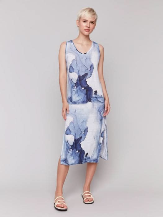 A person stands against a plain background wearing the Charlie B Sleeveless Linen Blend Dress with Side Slits in a blue abstract print, paired with white sandals—ideal for warm weather.