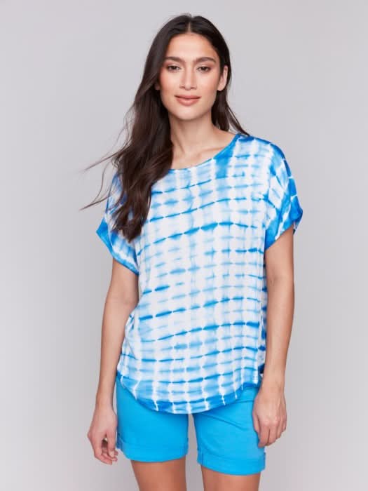 A woman faces forward in a Charlie B Tie Dye Dolman Short Sleeve Relaxed Top, blue and white, paired with blue shorts against a plain light gray background.