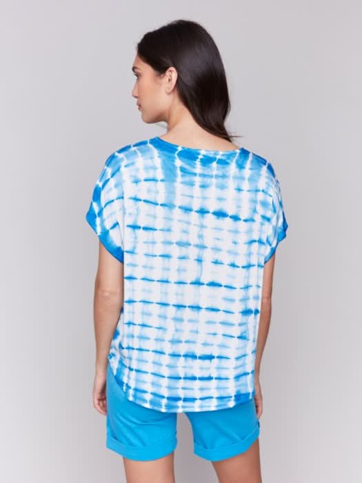 A woman faces forward in a Charlie B Tie Dye Dolman Short Sleeve Relaxed Top, blue and white, paired with blue shorts against a plain light gray background.
