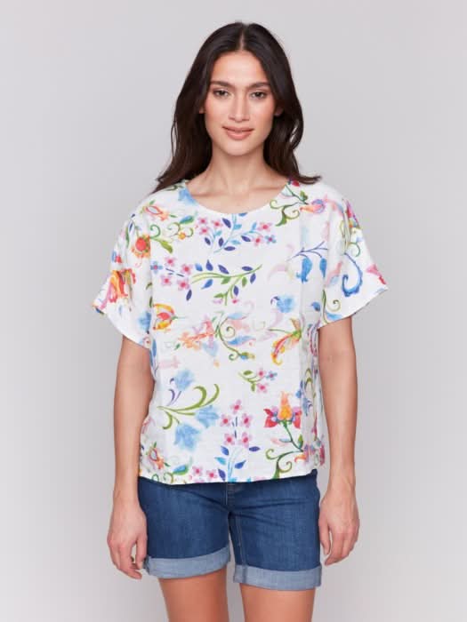 A woman stands against a plain background wearing the Charlie B Printed Linen Short Dolman Sleeve Top, light green cropped pants, and nude heeled sandals.