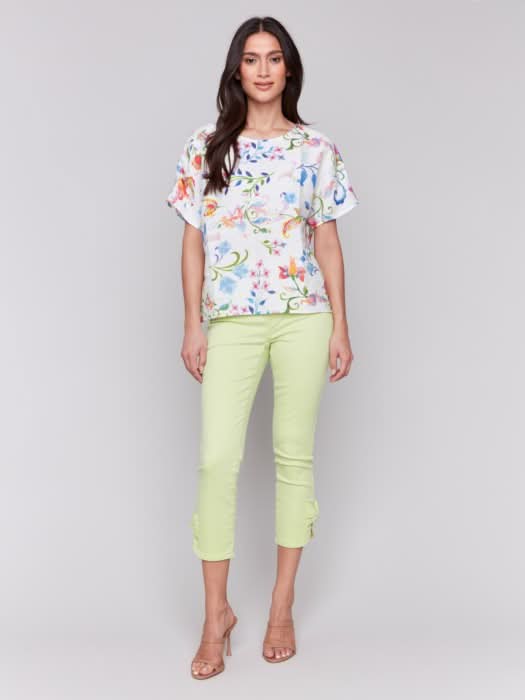 A woman stands against a plain background wearing the Charlie B Printed Linen Short Dolman Sleeve Top, light green cropped pants, and nude heeled sandals.