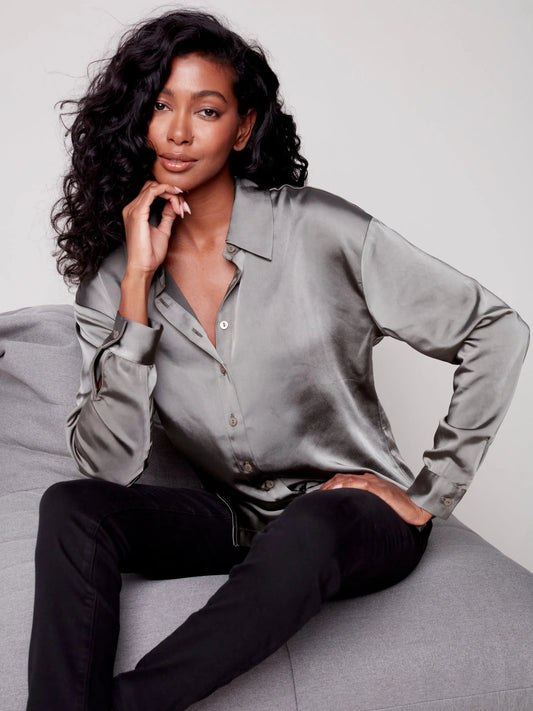 A stylish black woman is stylishly sitting on a couch in a Charlie B Satin Button Up Collard Shirt and black pants made of satin fabric.
