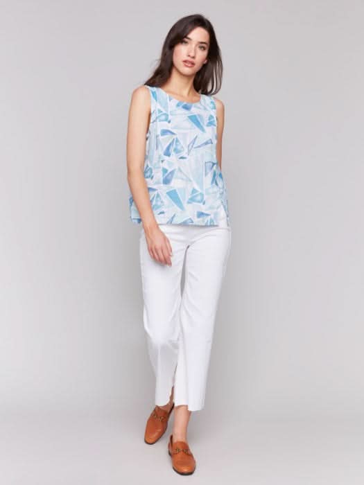 A woman models the Charlie B Sleeveless Linen Blend Top with Slit, featuring a geometric blue and white pattern. She pairs it with white pants against a light grey background. The breathable fabric provides all-day comfort.
