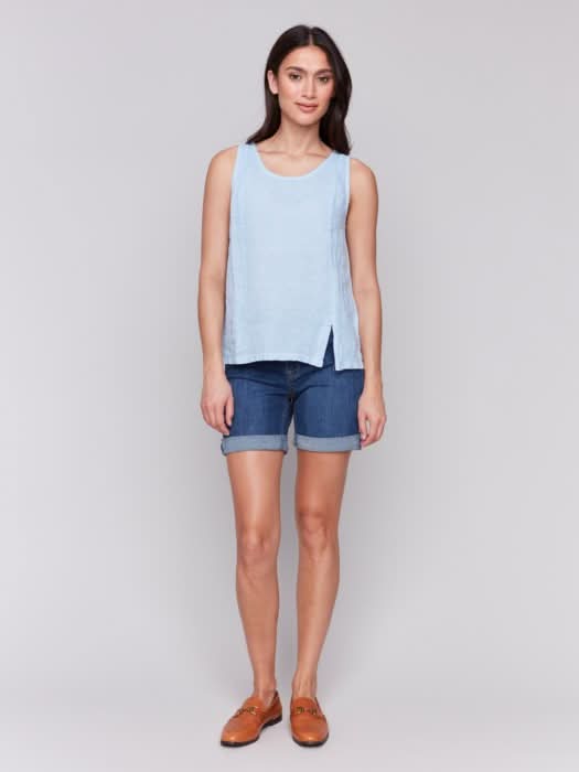 A fashionista with long dark hair models the Charlie B Sleeveless Linen Top with Slit in light blue paired with denim shorts, standing confidently against a plain gray background.