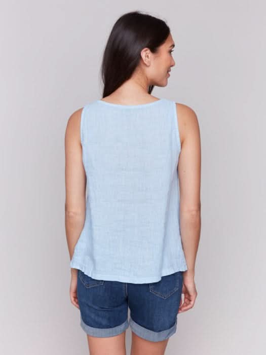 A fashionista with long dark hair models the Charlie B Sleeveless Linen Top with Slit in light blue paired with denim shorts, standing confidently against a plain gray background.