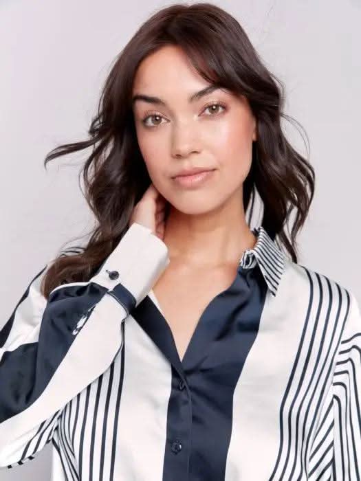 A woman wearing the Satin Twisted Detail Front Shirt by Charlie B along with jeans stands against a plain background. Her long brown hair complements the ensemble, giving her an elegant look, while she maintains a neutral expression.