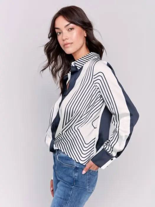 A woman wearing the Satin Twisted Detail Front Shirt by Charlie B along with jeans stands against a plain background. Her long brown hair complements the ensemble, giving her an elegant look, while she maintains a neutral expression.