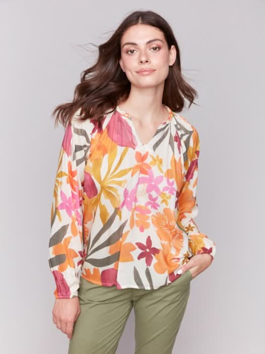 A person with long hair wearing a Charlie B Printed Viscose Long Sleeve Popover Blouse and green pants poses against a plain background, one hand in their pocket.