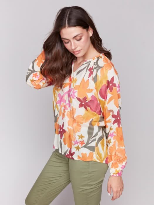 A person with long hair wearing a Charlie B Printed Viscose Long Sleeve Popover Blouse and green pants poses against a plain background, one hand in their pocket.