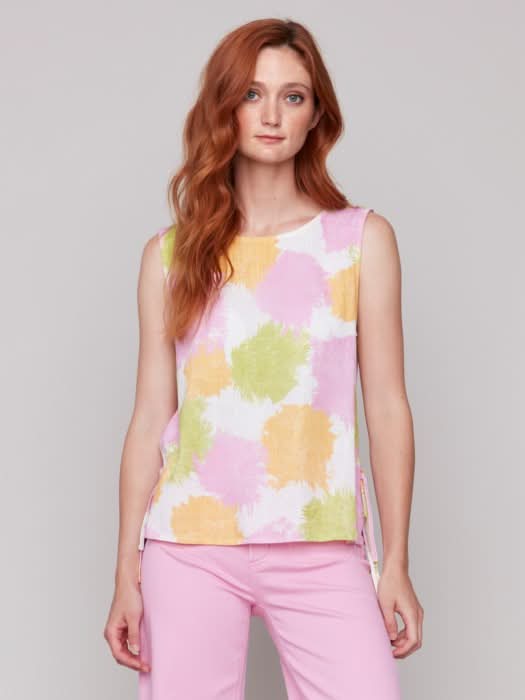 A woman with long red hair wears a Charlie B Sleeveless Linen Blend Top with Side Ties featuring pink, yellow, and green splotches, paired with light pink pants. She stands against a plain light background.