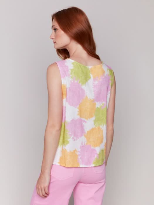 A woman with long red hair wears a Charlie B Sleeveless Linen Blend Top with Side Ties featuring pink, yellow, and green splotches, paired with light pink pants. She stands against a plain light background.