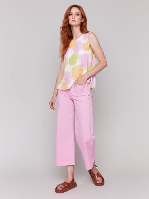 A woman with long red hair wears a Charlie B Sleeveless Linen Blend Top with Side Ties featuring pink, yellow, and green splotches, paired with light pink pants. She stands against a plain light background.