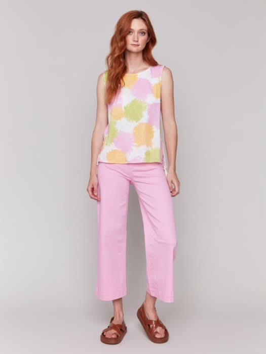 A woman with long red hair wears a Charlie B Sleeveless Linen Blend Top with Side Ties featuring pink, yellow, and green splotches, paired with light pink pants. She stands against a plain light background.