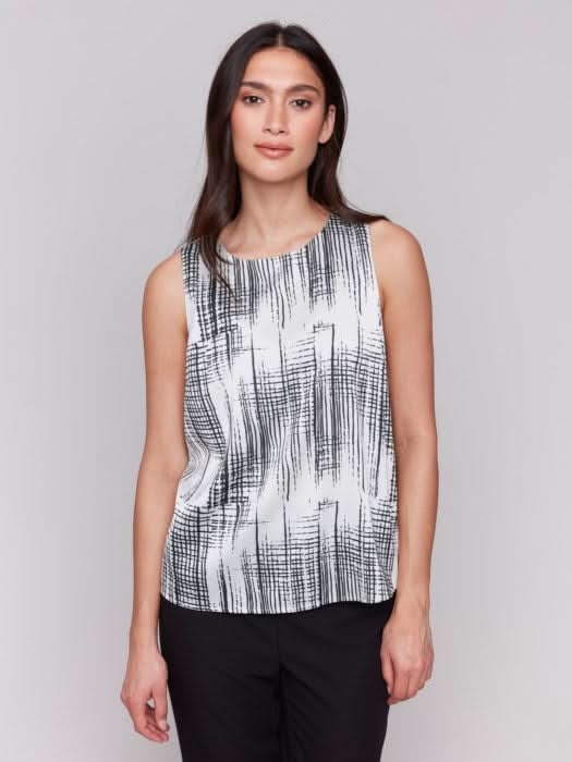 A woman with long hair models the Charlie B Printed Satin Sleeveless Top—stylishly bra-friendly and black-and-white printed—paired with black pants, set against a plain gray background.