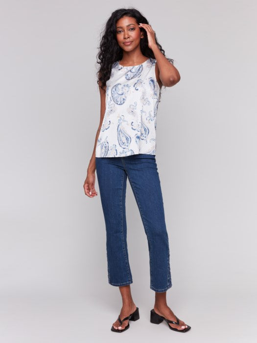 Woman with long wavy hair wears the Charlie B Printed Satin Sleeveless Top featuring blue paisley patterns, paired with blue jeans, standing against a plain light background.