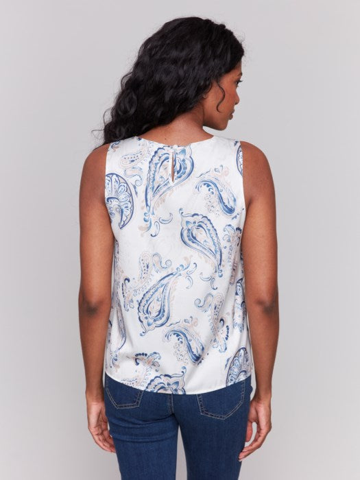 Woman with long wavy hair wears the Charlie B Printed Satin Sleeveless Top featuring blue paisley patterns, paired with blue jeans, standing against a plain light background.