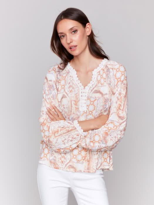 A woman poses with arms crossed in a Charlie B Raglan Blouse with a V-notch neckline and intricate pastel embroidery, paired with white pants, adding elegance to her ensemble.