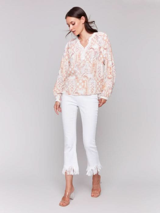 A woman poses with arms crossed in a Charlie B Raglan Blouse with a V-notch neckline and intricate pastel embroidery, paired with white pants, adding elegance to her ensemble.