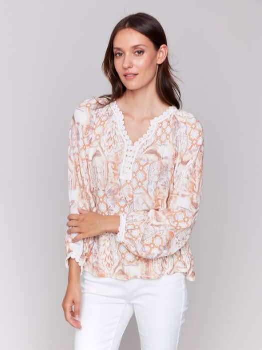 A woman poses with arms crossed in a Charlie B Raglan Blouse with a V-notch neckline and intricate pastel embroidery, paired with white pants, adding elegance to her ensemble.