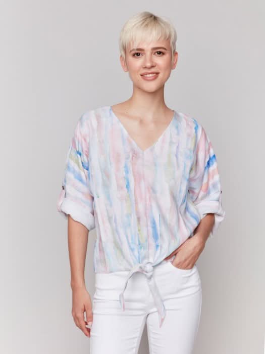 A person stands against a neutral background wearing the Charlie B 3/4 Rolled Cuff Sleeve V Neck in pastel tie-dye, paired with white cropped pants and sandals, one hand in their pocket for a relaxed, casual vibe.