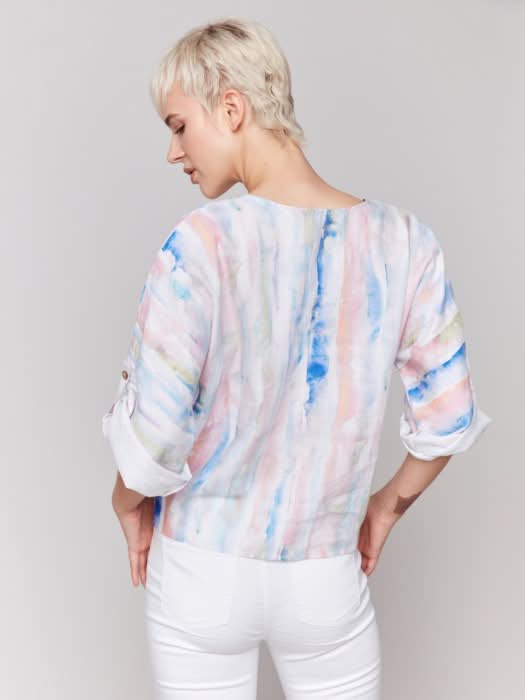 A person stands against a neutral background wearing the Charlie B 3/4 Rolled Cuff Sleeve V Neck in pastel tie-dye, paired with white cropped pants and sandals, one hand in their pocket for a relaxed, casual vibe.