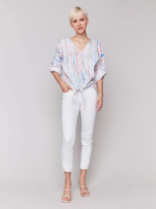 A person stands against a neutral background wearing the Charlie B 3/4 Rolled Cuff Sleeve V Neck in pastel tie-dye, paired with white cropped pants and sandals, one hand in their pocket for a relaxed, casual vibe.