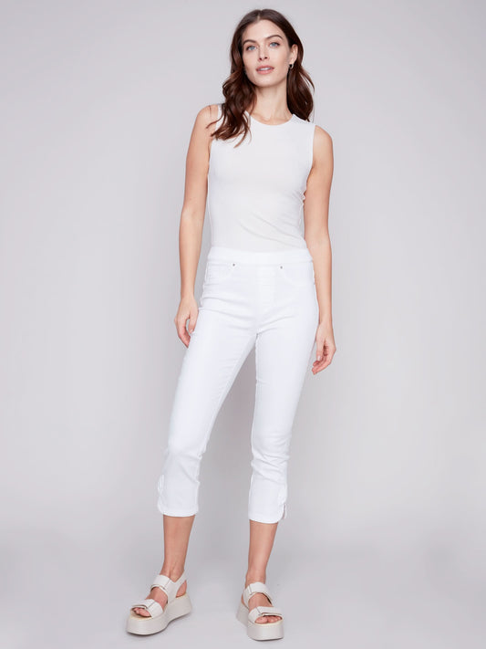 A woman stands against a plain background, wearing Charlie B's Pull-On Jeans With Bow Detail, paired with a sleeveless white top and white platform sandals for effortless summer charm.