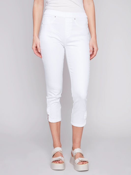 A woman stands against a plain background, wearing Charlie B's Pull-On Jeans With Bow Detail, paired with a sleeveless white top and white platform sandals for effortless summer charm.