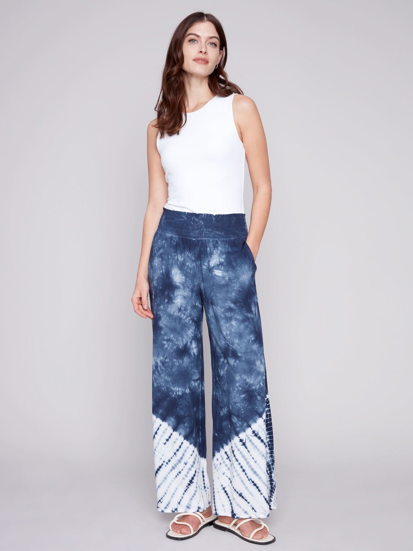A person wearing Charlie B's Gradient Palazzo Pant in blue and white tie-dye, made of flowy fabric, paired with white sandals, standing against a gray background.