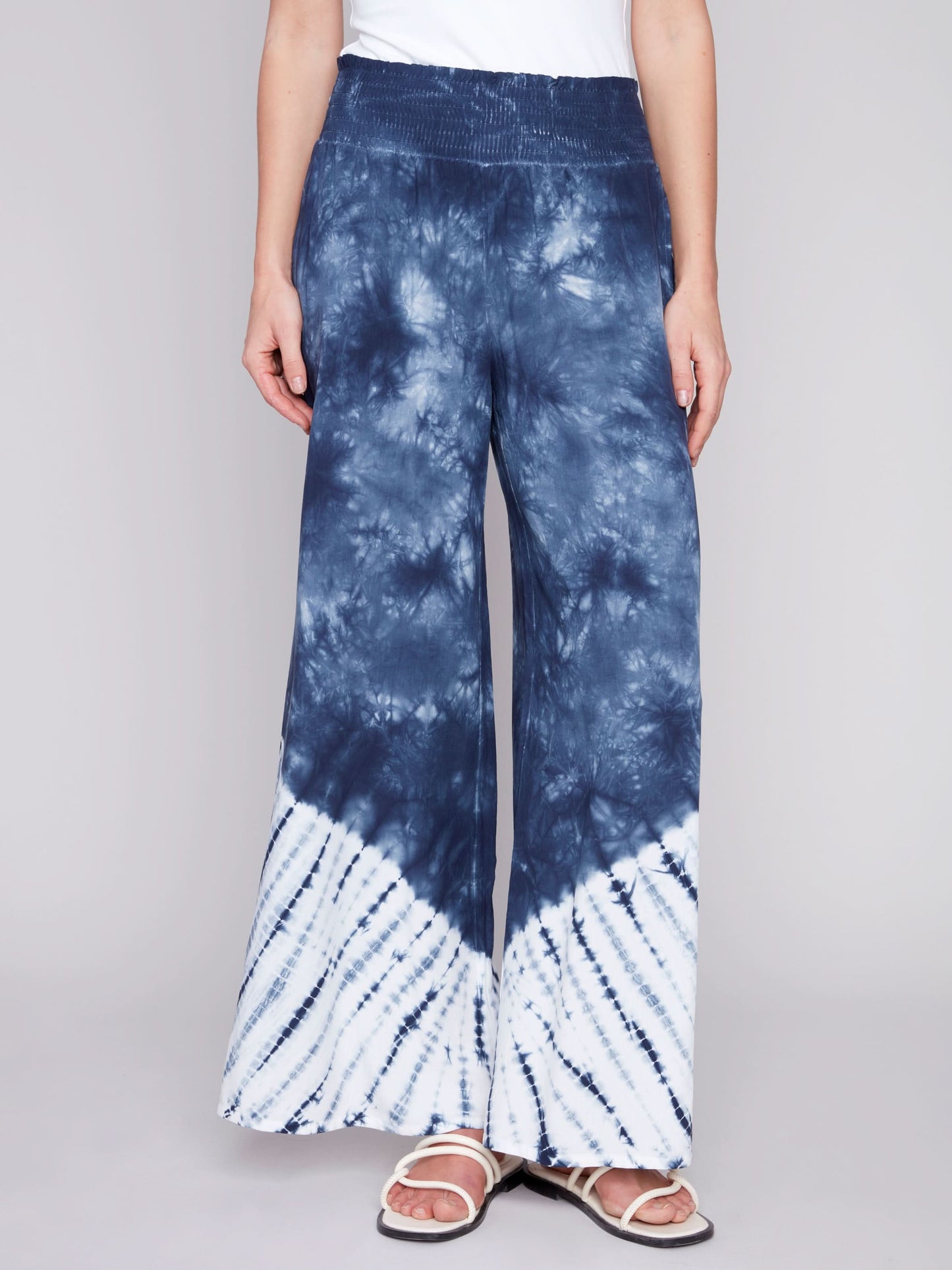 A person wearing Charlie B's Gradient Palazzo Pant in blue and white tie-dye, made of flowy fabric, paired with white sandals, standing against a gray background.