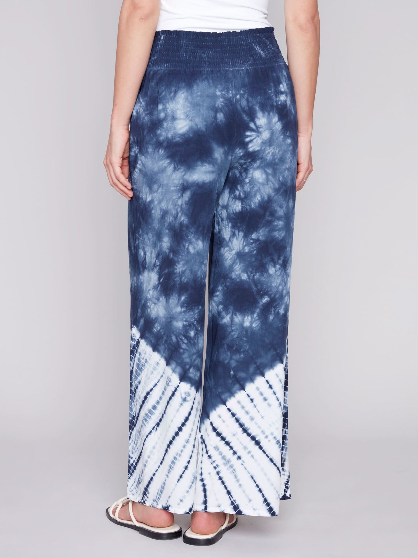 A person wearing Charlie B's Gradient Palazzo Pant in blue and white tie-dye, made of flowy fabric, paired with white sandals, standing against a gray background.