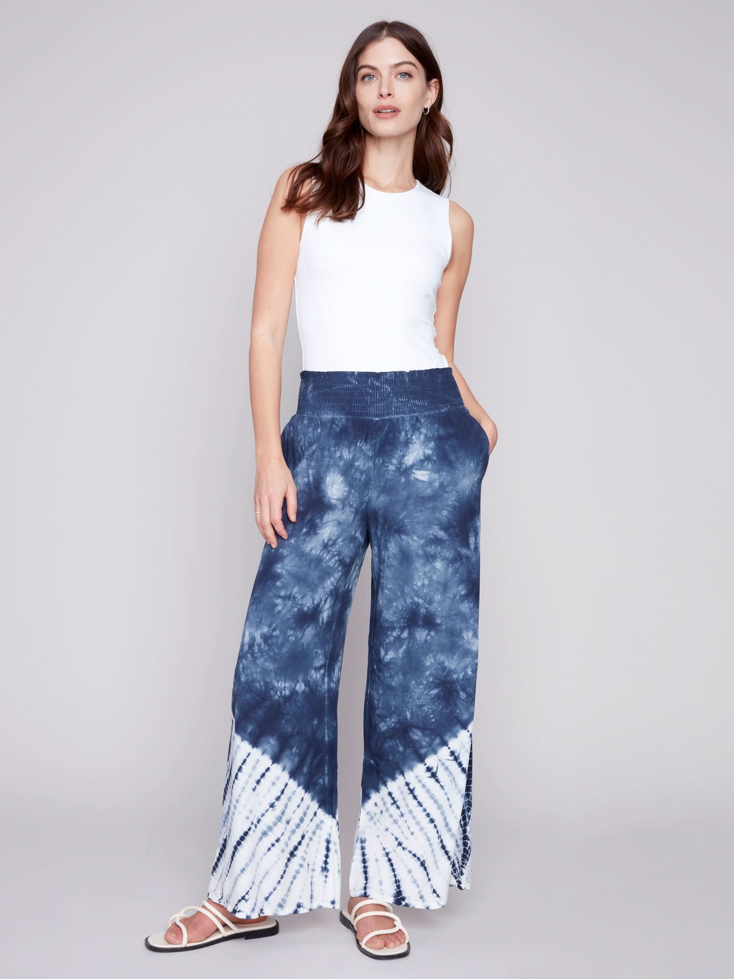 A person wearing Charlie B's Gradient Palazzo Pant in blue and white tie-dye, made of flowy fabric, paired with white sandals, standing against a gray background.