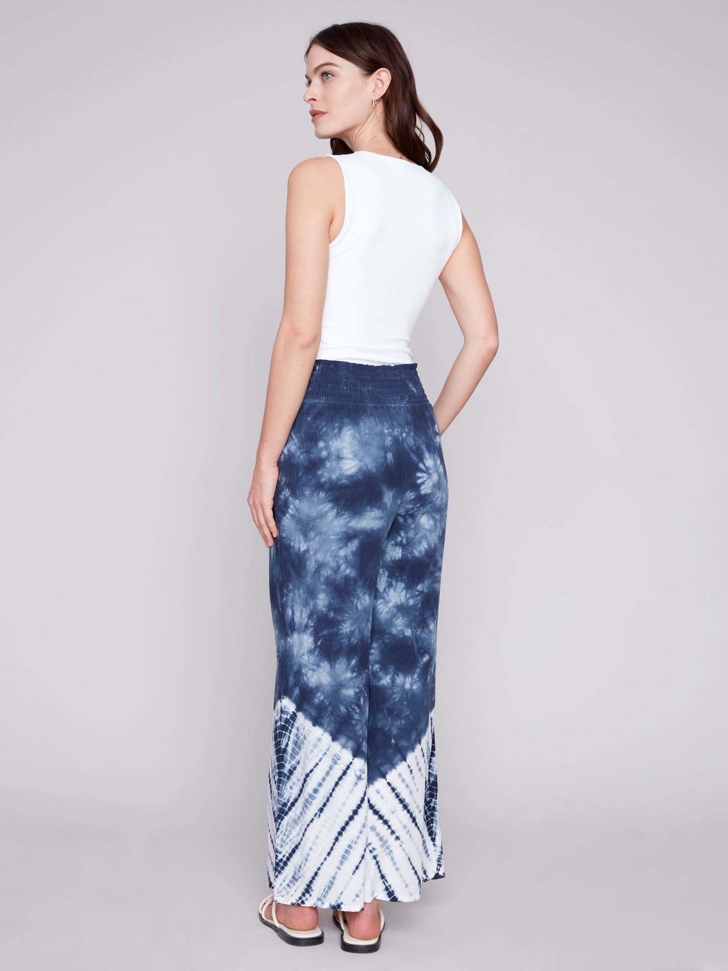 A person wearing Charlie B's Gradient Palazzo Pant in blue and white tie-dye, made of flowy fabric, paired with white sandals, standing against a gray background.