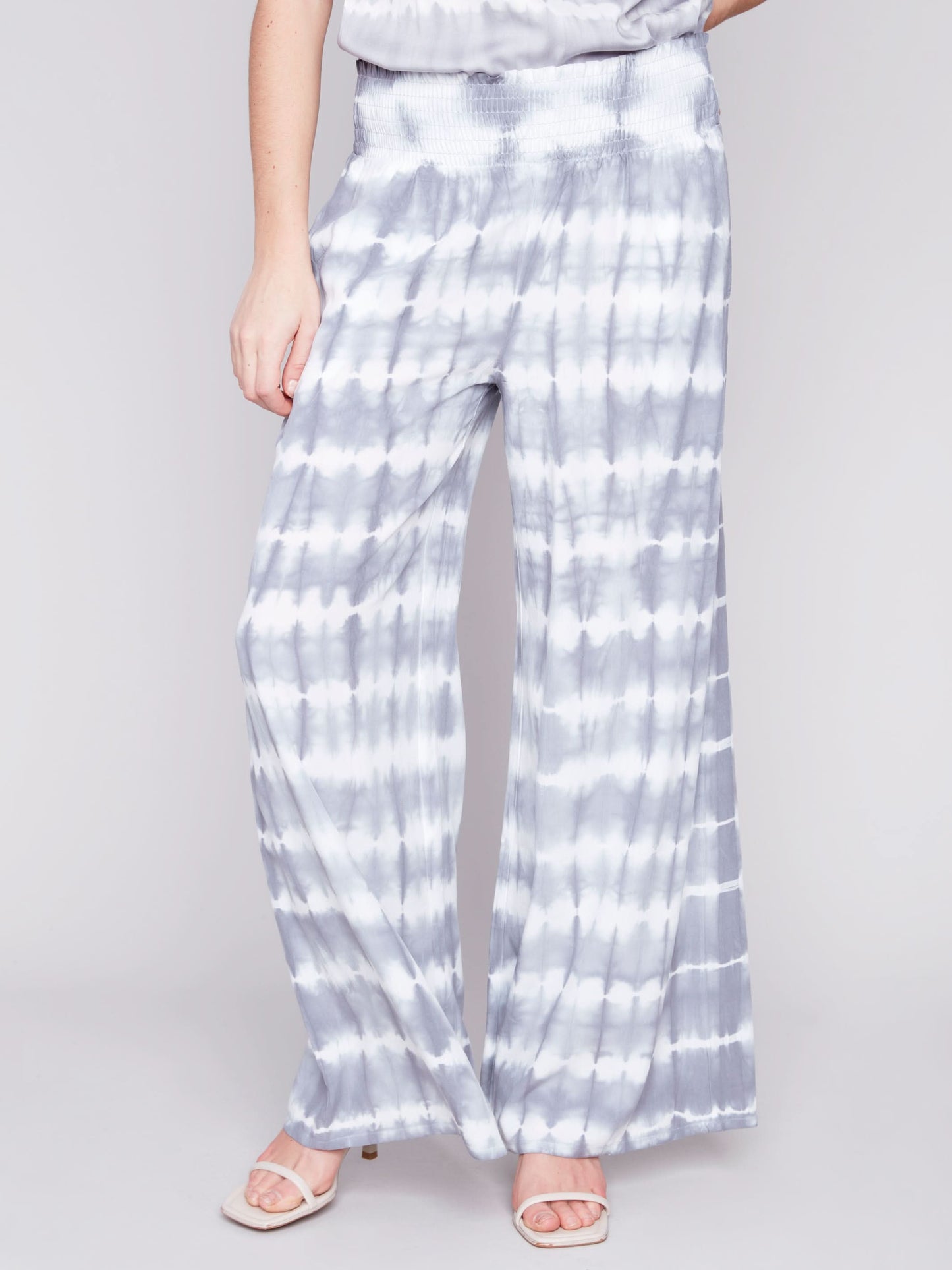 A person wearing Charlie B's Gradient Palazzo Pant in blue and white tie-dye, made of flowy fabric, paired with white sandals, standing against a gray background.