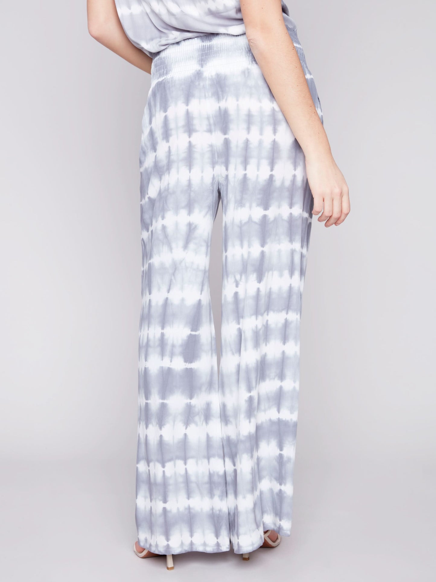 A person wearing Charlie B's Gradient Palazzo Pant in blue and white tie-dye, made of flowy fabric, paired with white sandals, standing against a gray background.