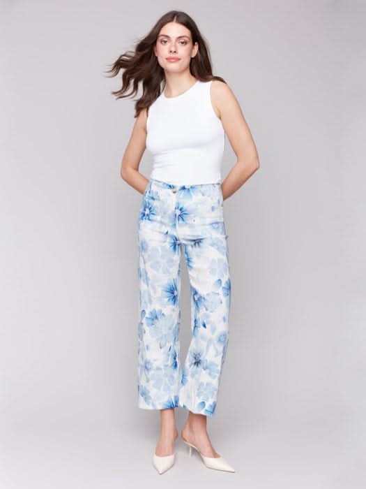 A person is wearing Charlie B's Printed Cropped Linen Blend Pants with a black floral pattern, a black top, and beige open-toe slides while standing against a plain background.