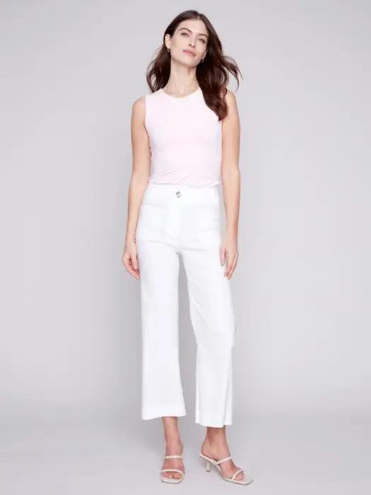 A woman standing against a gray backdrop, wearing a pink sleeveless top and white Charlie B cropped straight leg pants, paired with white heeled sandals.