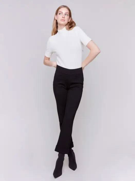 Dressed in stylish Cuffed Ponte Pants by Charlie B and chic black heeled boots, an individual stands with one hand in their pocket, radiating a fashion-forward aura against a plain background.