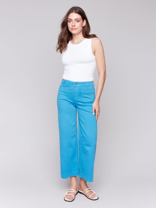 A woman stands against a plain background wearing a white sleeveless top, Charlie B Patch Pocket Flare Twill Pants in bright blue, and white sandals.