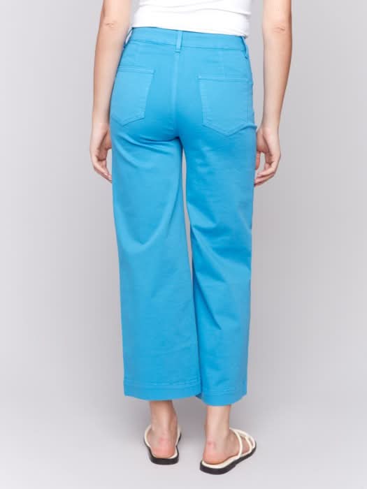 A woman stands against a plain background wearing a white sleeveless top, Charlie B Patch Pocket Flare Twill Pants in bright blue, and white sandals.