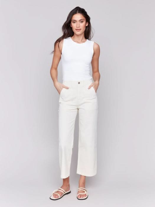 A person in a white sleeveless top and light-colored Cropped Utility Pocket Twill Pants by Charlie B stands against a plain gray background. Their hands rest casually in their pockets, complemented by white sandals.