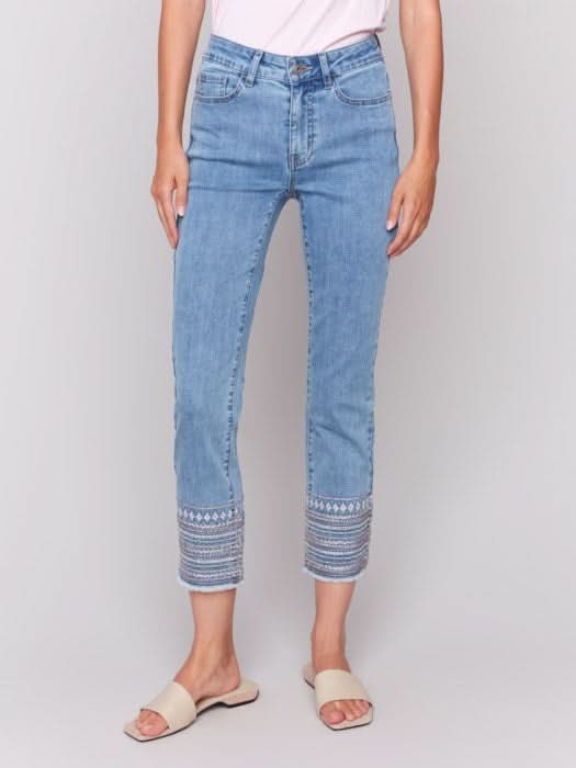 A woman stands against a plain background wearing a sleeveless white top, Charlie B Embroidered Hem Jeans in a slim leg silhouette, and beige sandals, with one hand in her pocket.