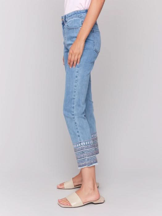 A woman stands against a plain background wearing a sleeveless white top, Charlie B Embroidered Hem Jeans in a slim leg silhouette, and beige sandals, with one hand in her pocket.
